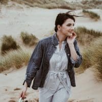 woman on beach wearing denim jacket and striped dress with shoes in hand
